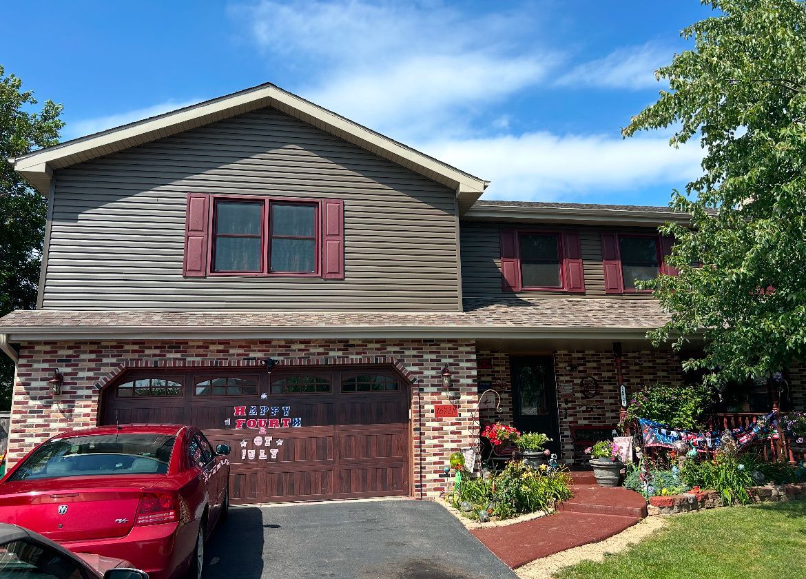 Two-story brick house with red shutters, a red car in the driveway, and a garden in front.