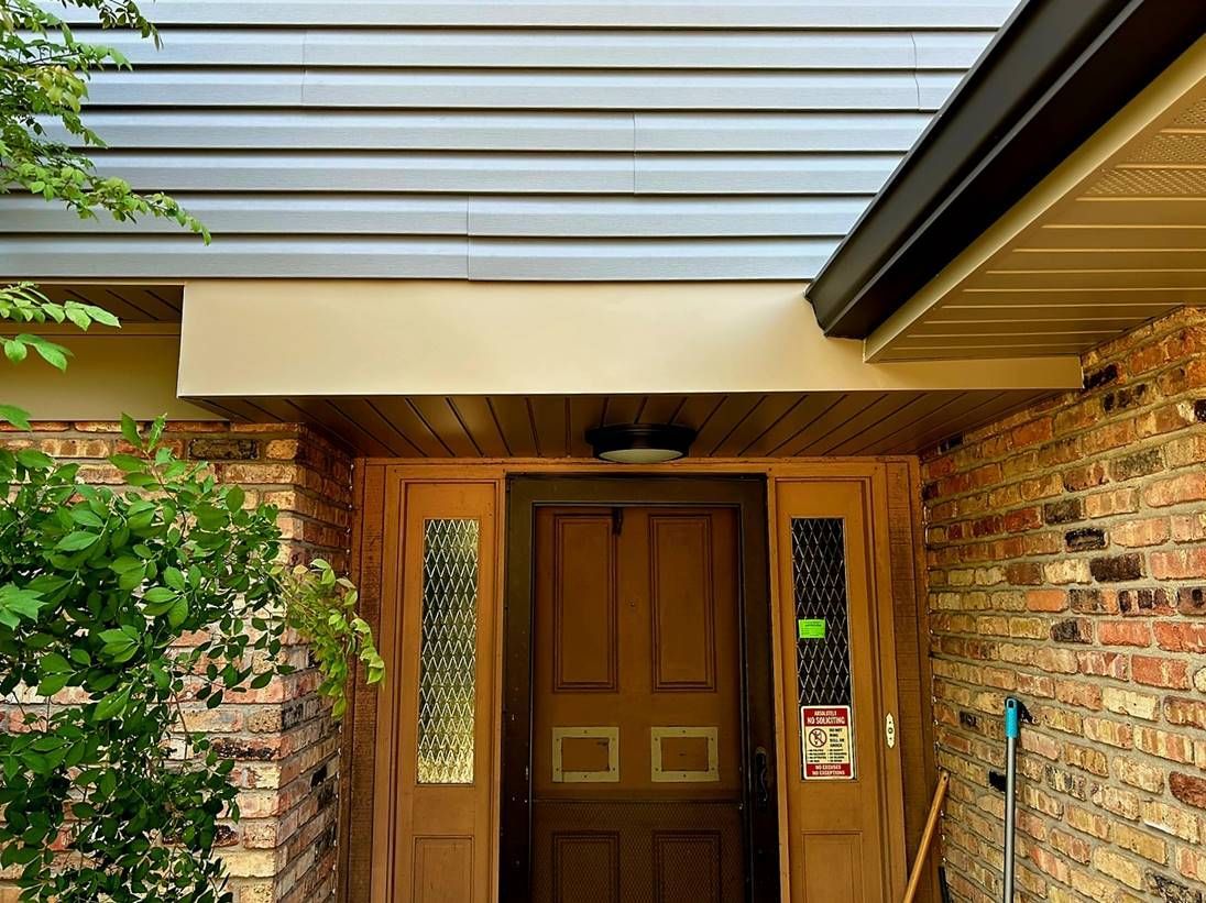 Front door under a porch roof, framed by brick walls and a wooden trim, with a leafy plant on the left.
