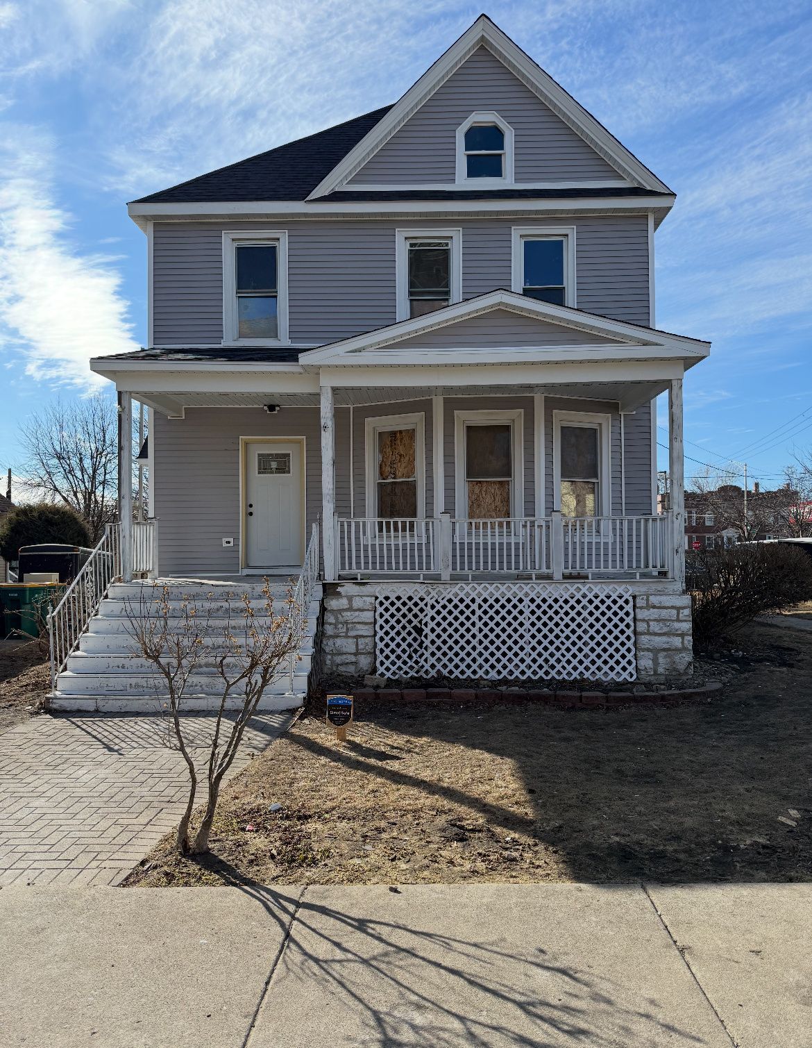Front view of a weathered two-story house with a porch, steps, and a driveway under a blue sky