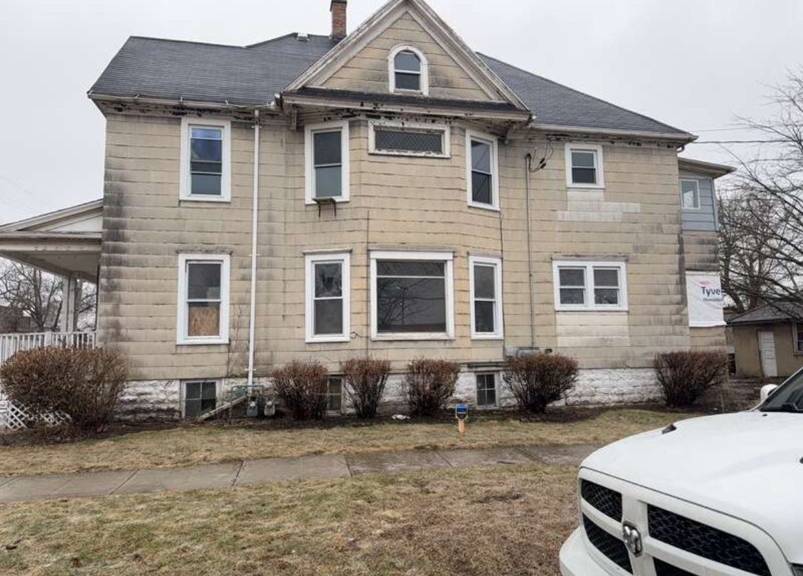 Two-story beige house with a gray roof and white car parked in front on a cloudy day