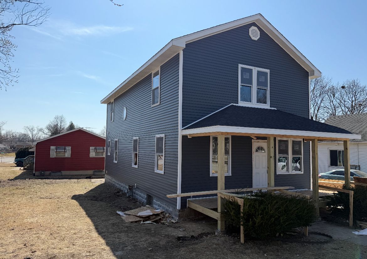 Blue two-story house with white trim and a small porch beside a red shed in a grassy yard