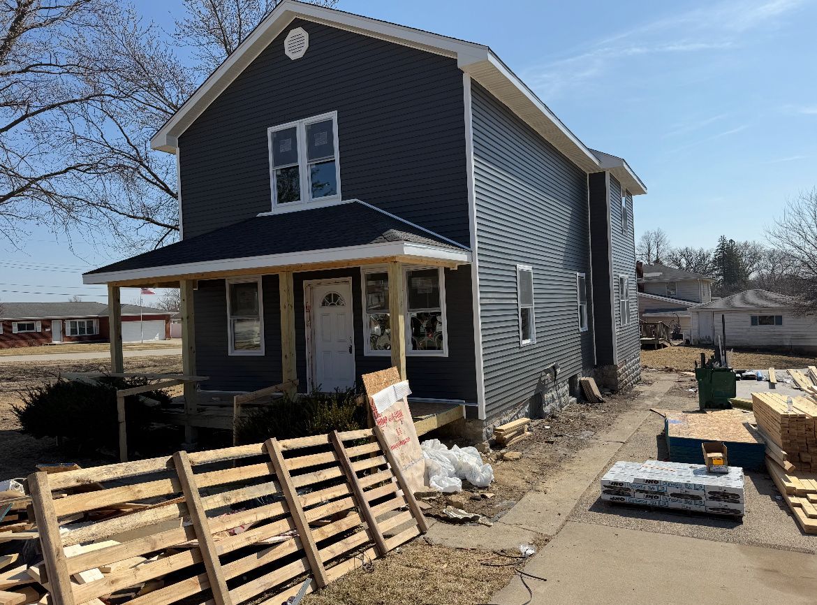 Two-story dark gray house under construction with porch, pallets, and building materials in front yard