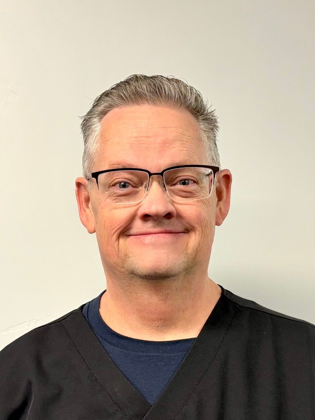 A person wearing glasses and a dark V-neck shirt over a navy sweater, smiling against a plain, light-colored background.