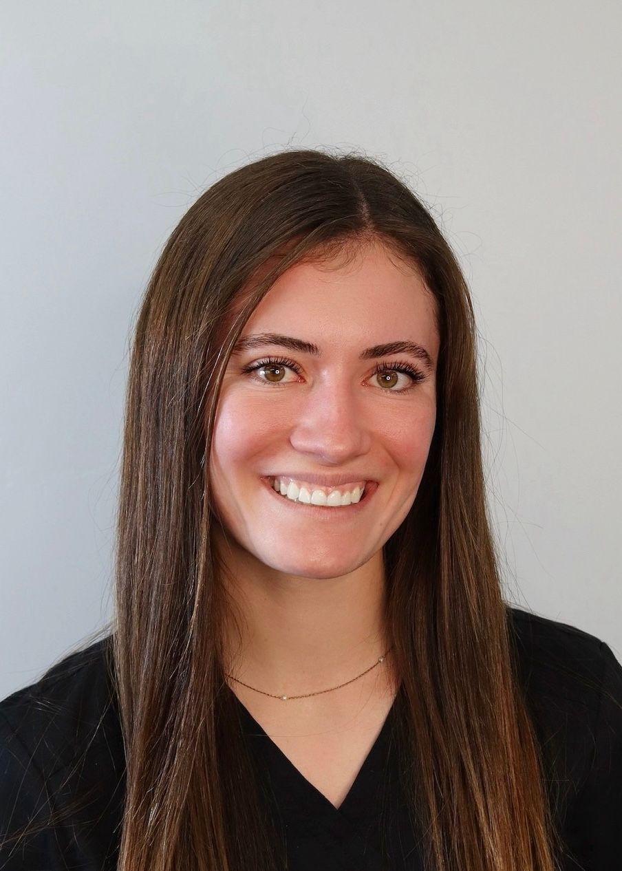 A headshot of a smiling person with long brown hair, wearing a black shirt, set against a plain light gray background.