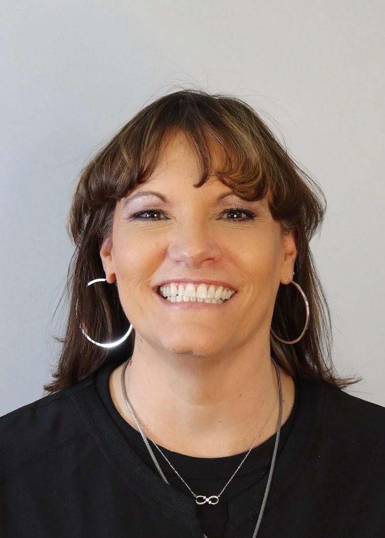 A person with brown hair and hoop earrings smiling at the camera against a plain, light gray background.