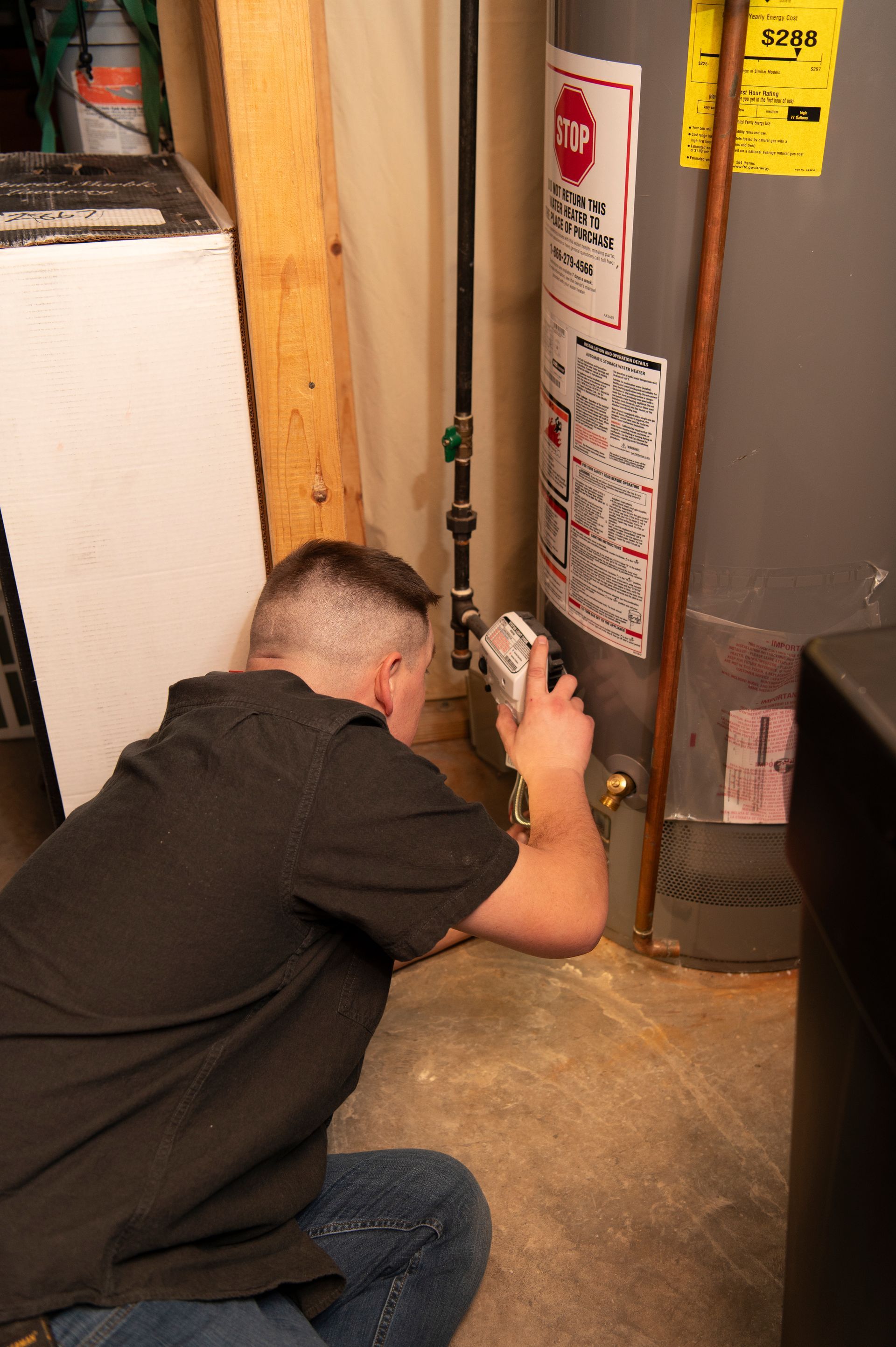 A man is kneeling down in front of a water heater in a basement.