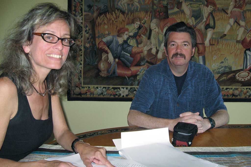 Two people seated at a table with papers, smiling in a room with a colorful wall tapestry.