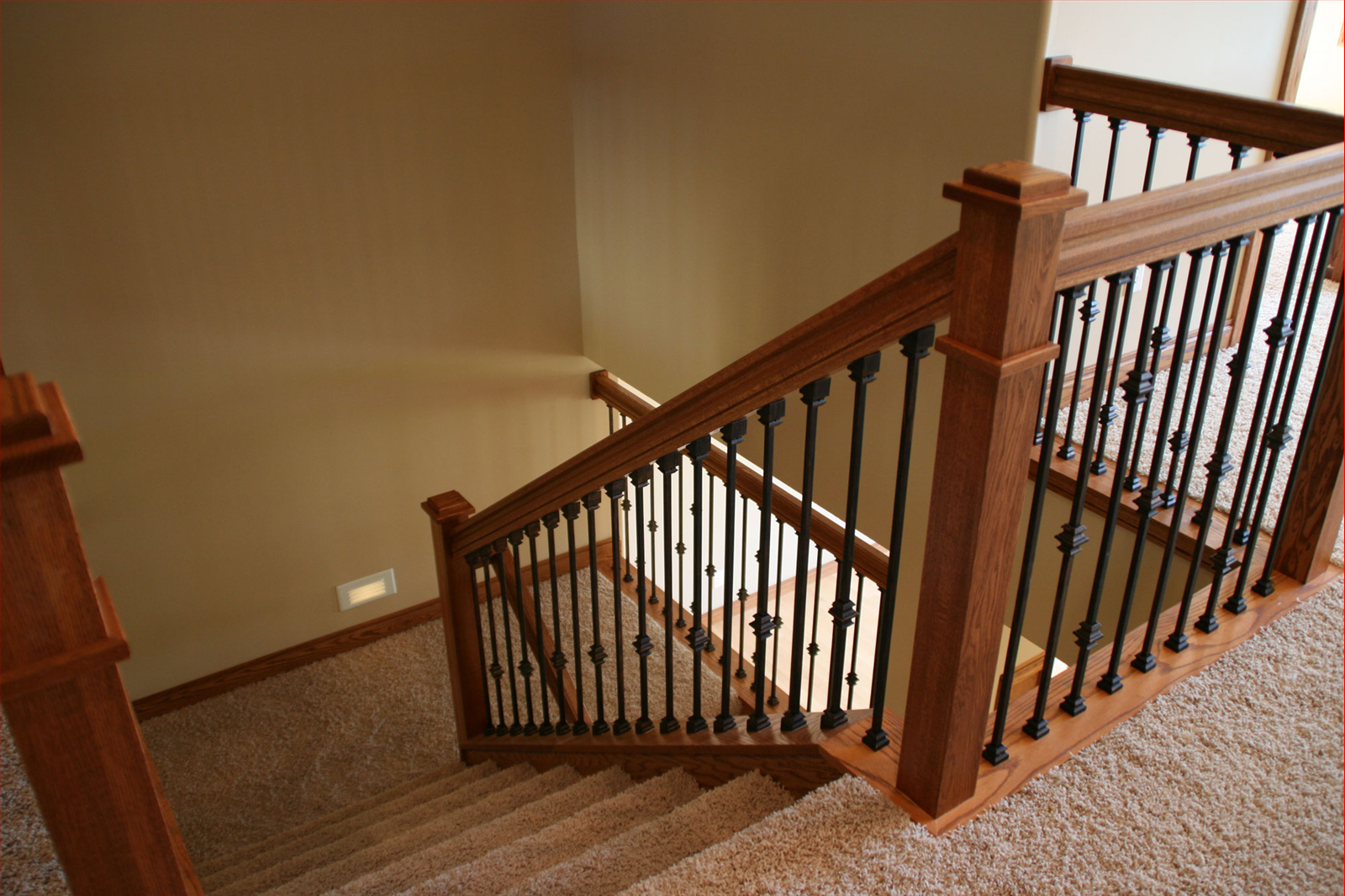 Wooden staircase with black metal balusters, tan walls, and carpeted steps.