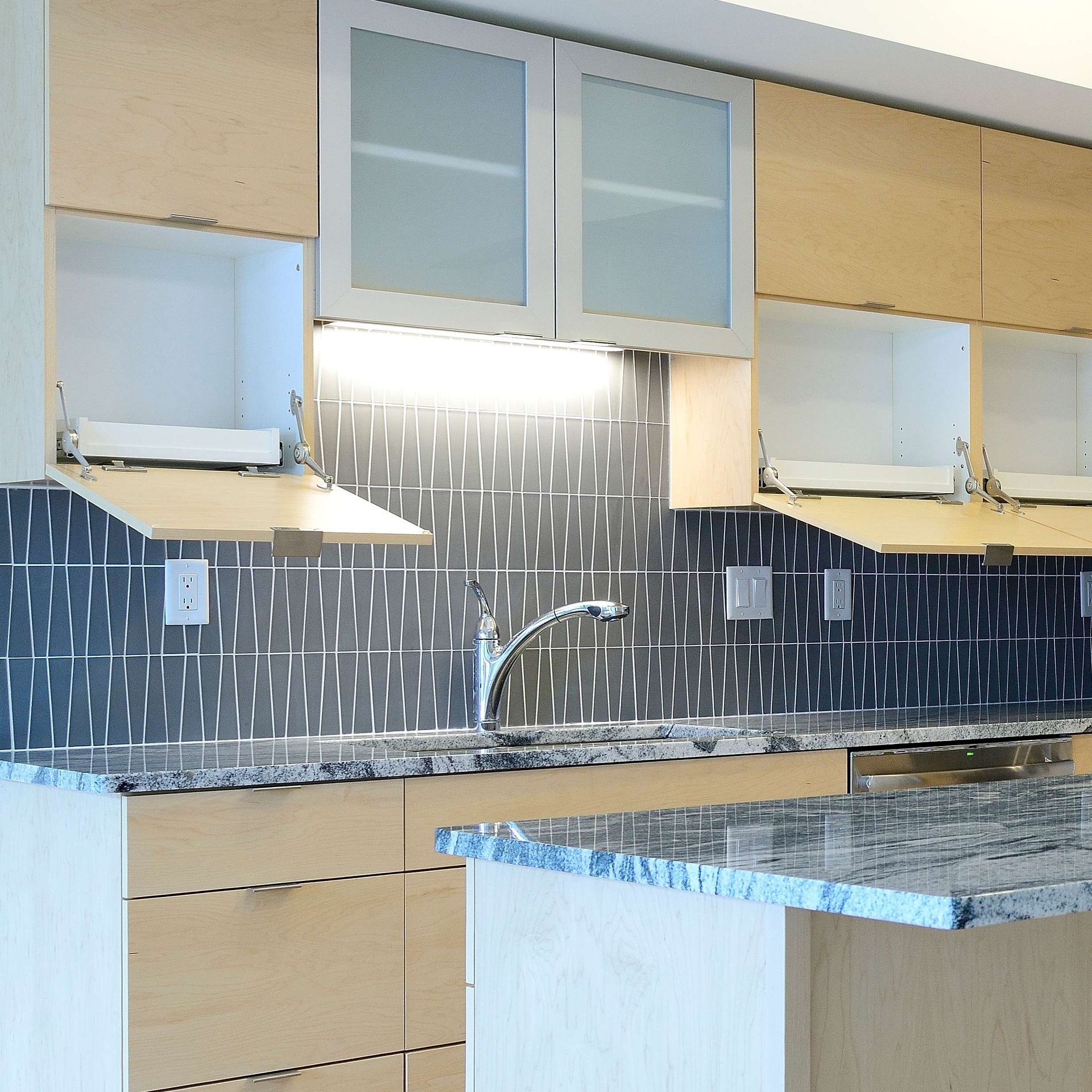 Modern kitchen with light wood cabinets, gray countertops, and blue tile backsplash.
