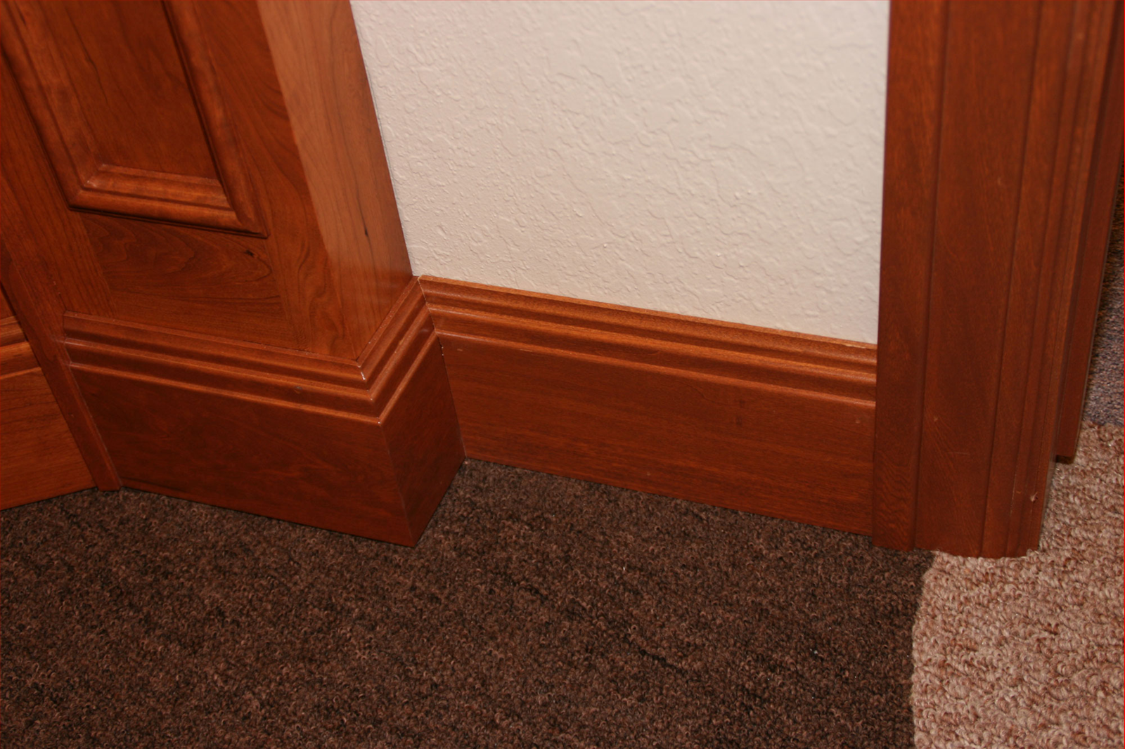Close-up of brown wooden door trim and baseboard against a white wall and brown carpet.