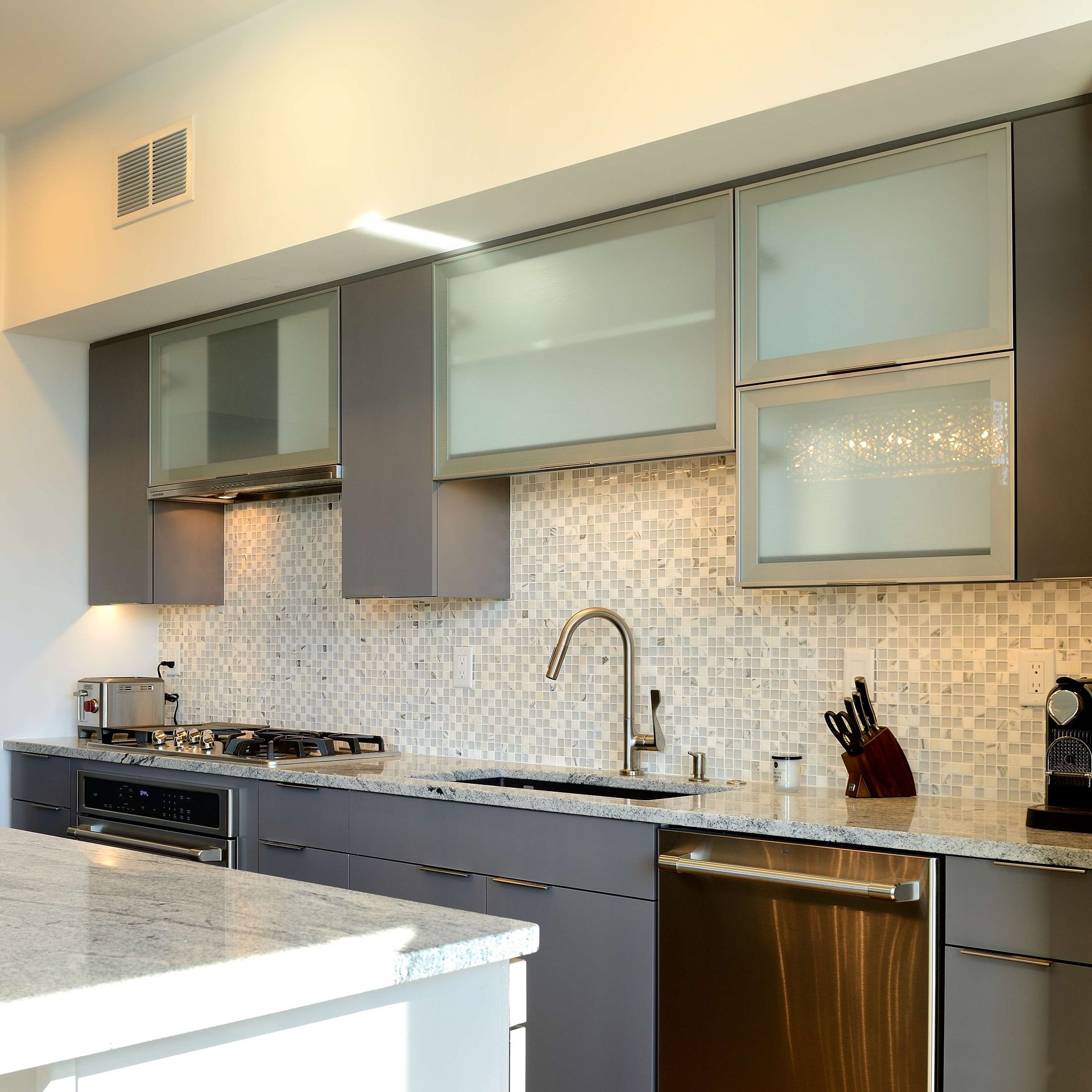 Modern kitchen with gray cabinets, frosted glass doors, stainless steel appliances, and a mosaic backsplash.