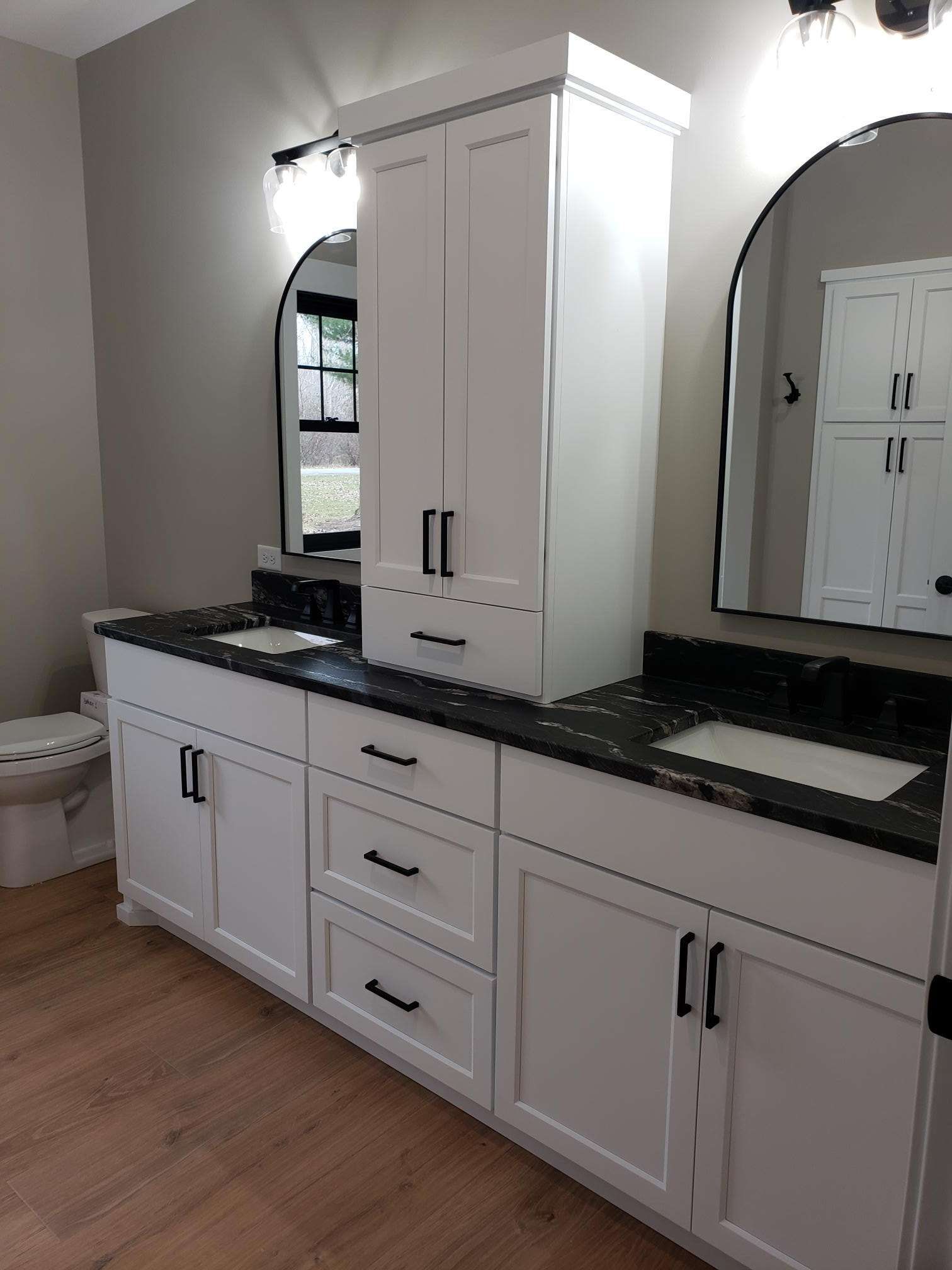 Bathroom with white cabinets, black countertops, mirrors, and a tall storage unit.