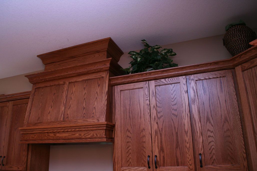 Wooden kitchen cabinets with a range hood, topped with greenery, against a white ceiling.