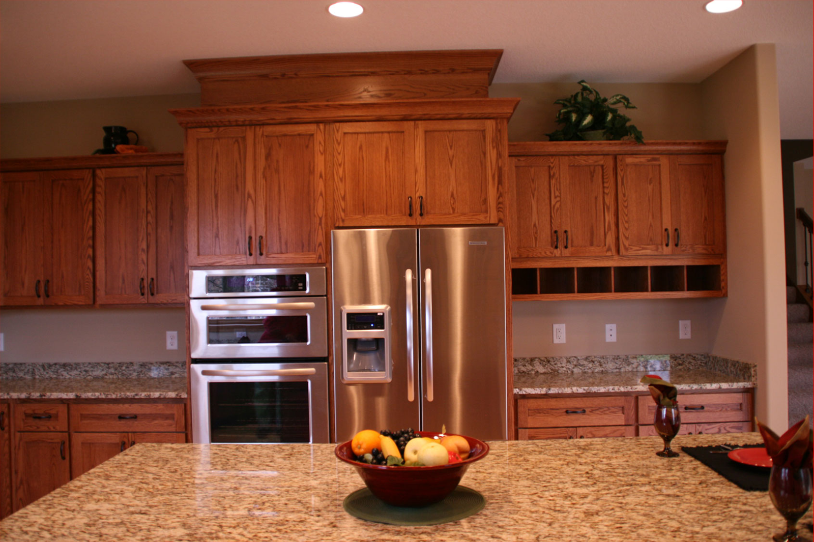 Kitchen with stainless steel refrigerator and oven, wooden cabinets, granite countertops, and island with bowl of fruit.
