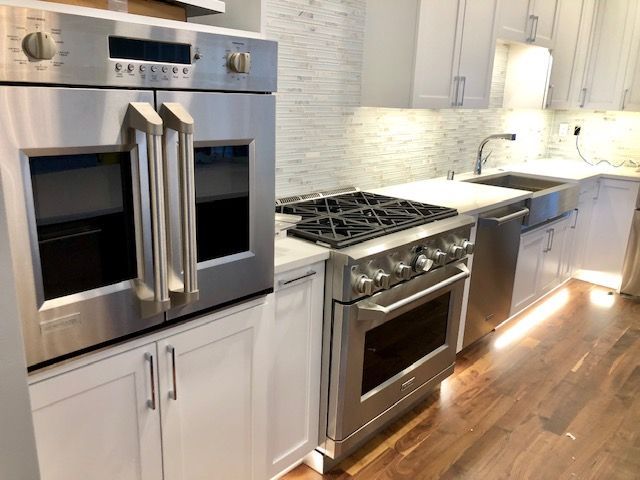 Modern kitchen with stainless steel appliances: oven, range, and sink. White cabinets, wood floor, and backsplash.