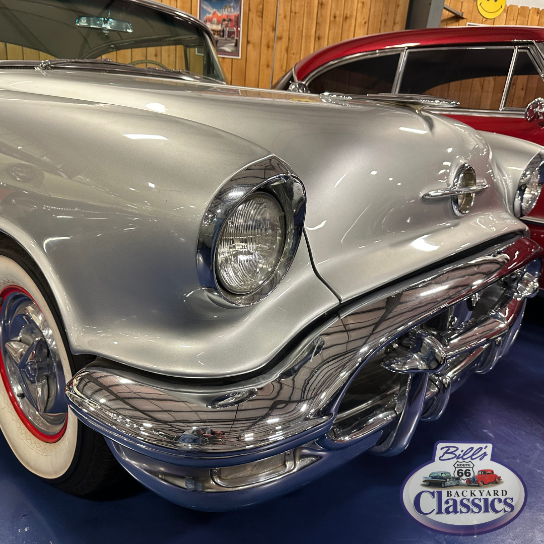 Silver vintage car with chrome accents and white-wall tires, red car in background.