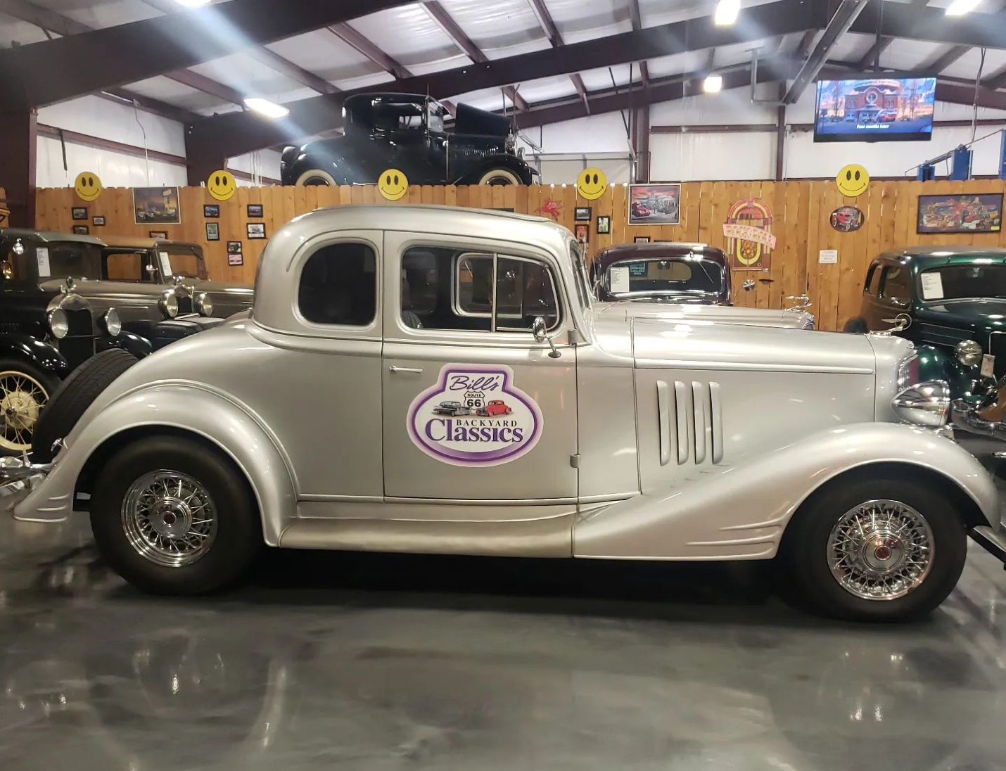 Silver classic car parked inside a large showroom with other vintage vehicles.