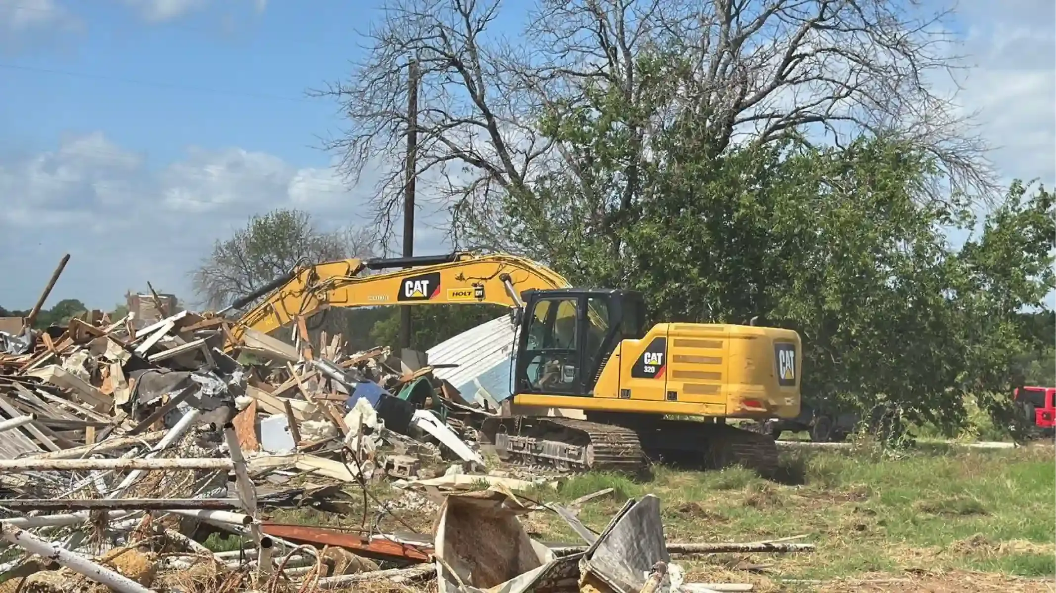 Yellow excavator clearing rubble and debris in a demolition site with trees in the background