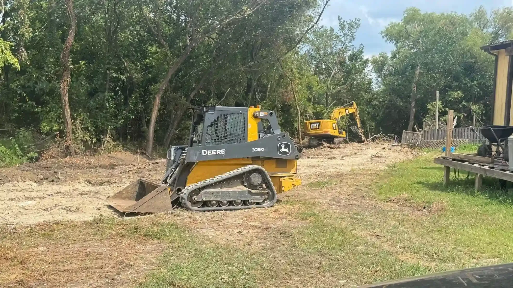 Yellow and gray skid-steer clearing brush beside a wooded path, with another excavator in the background