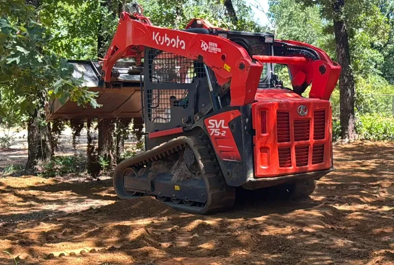 Red Kubota compact track loader parked on dirt in a wooded area.