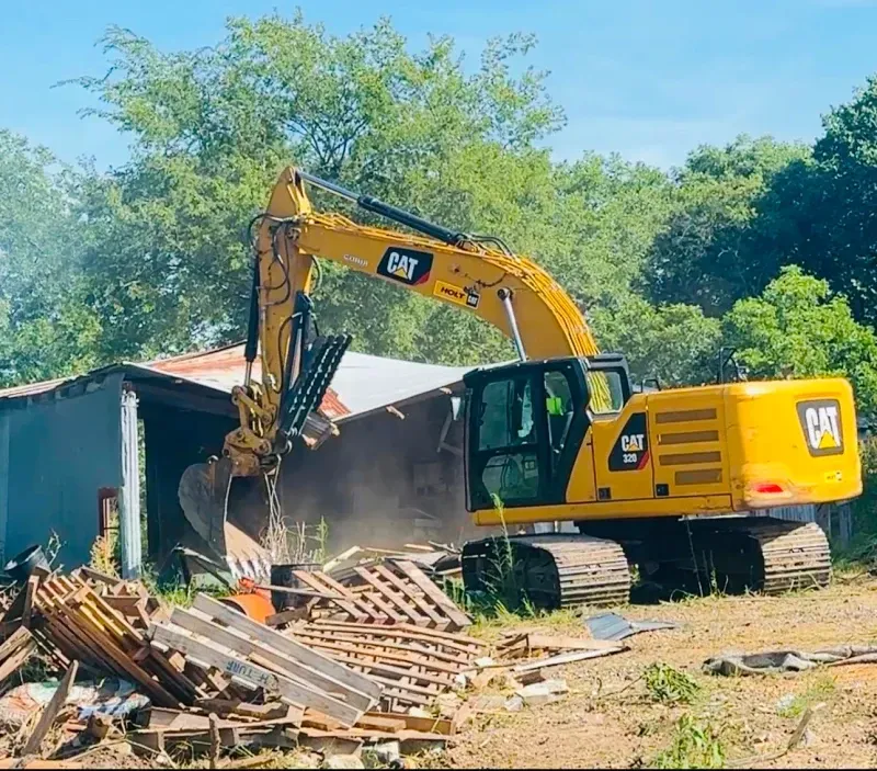 Yellow excavator demolishing a small building amid debris and dust outdoors