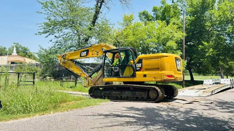Yellow excavator on a roadside, with a worker in the cab beside trees and a fence.