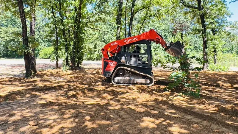 Orange excavator clearing dirt in a wooded area with sunlight filtering through trees