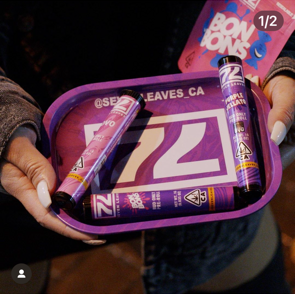 A person holding a purple tray with cannabis vape pens and candy.