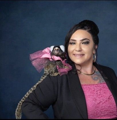 Woman in pink top and black blazer, with monkey wearing a pink tutu on her shoulder, dark blue background