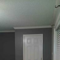 A white door set in a gray wall under a white textured ceiling with crown molding and a window with blinds on the right.
