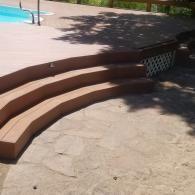Curved wooden steps lead up to a swimming pool deck, with a stone patio and white lattice skirting in the background.