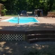 A rectangular swimming pool set in a backyard with a wooden deck, lattice skirting, and a car parked in the background.
