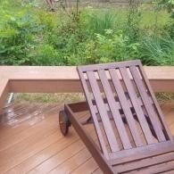 A brown wooden lounge chair sits on a deck overlooking a lush green garden.