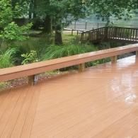 A wooden deck with a built-in bench overlooks a lush backyard with a small bridge and trees.