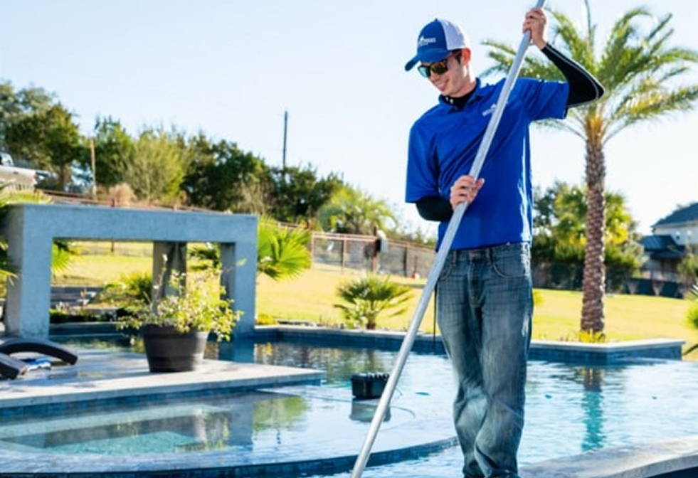A pool technician in a blue uniform cleans an outdoor swimming pool on a sunny day.