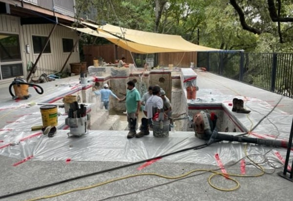 Workers in casual attire renovate a pool area covered with plastic sheets under a large shade sail in an outdoor setting.