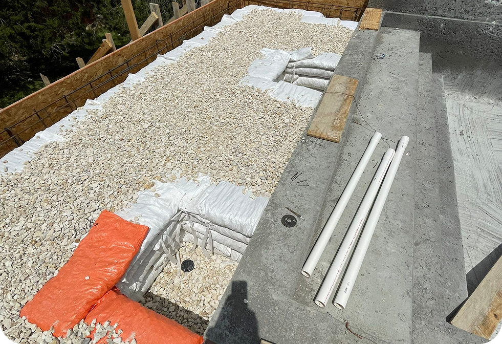 Construction site showing gravel, white sandbags, and PVC pipes arranged near concrete steps at a pool renovation.