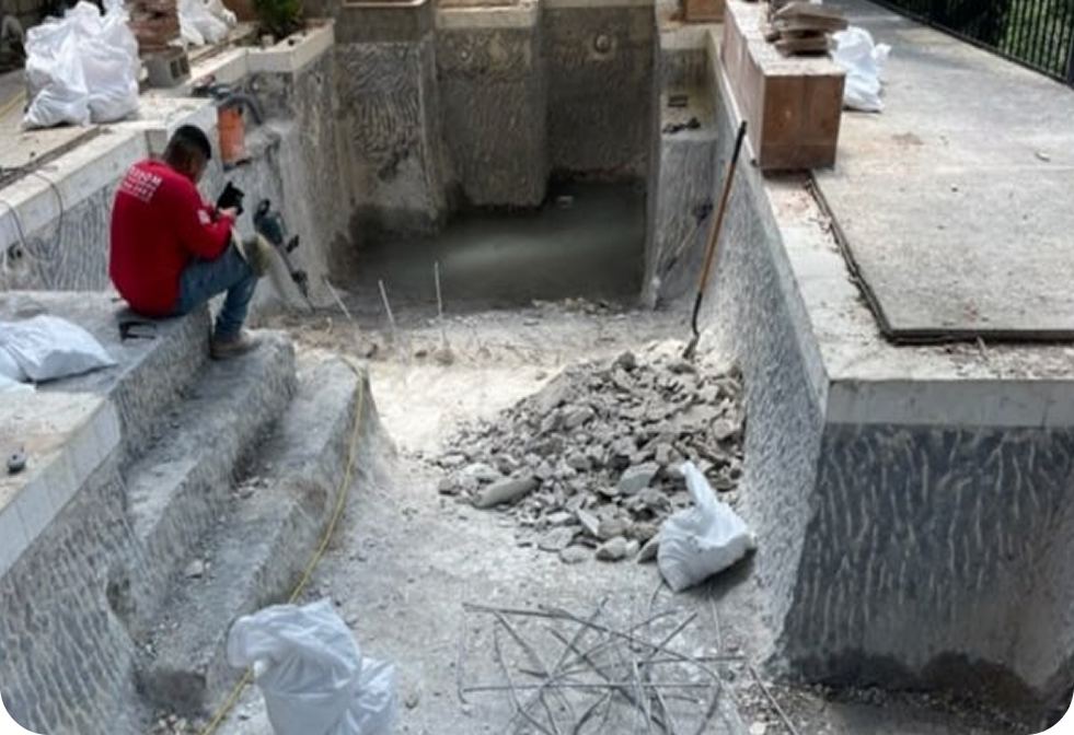 A person in a red shirt sits on the stairs of an empty swimming pool undergoing renovation, surrounded by debris and tools.