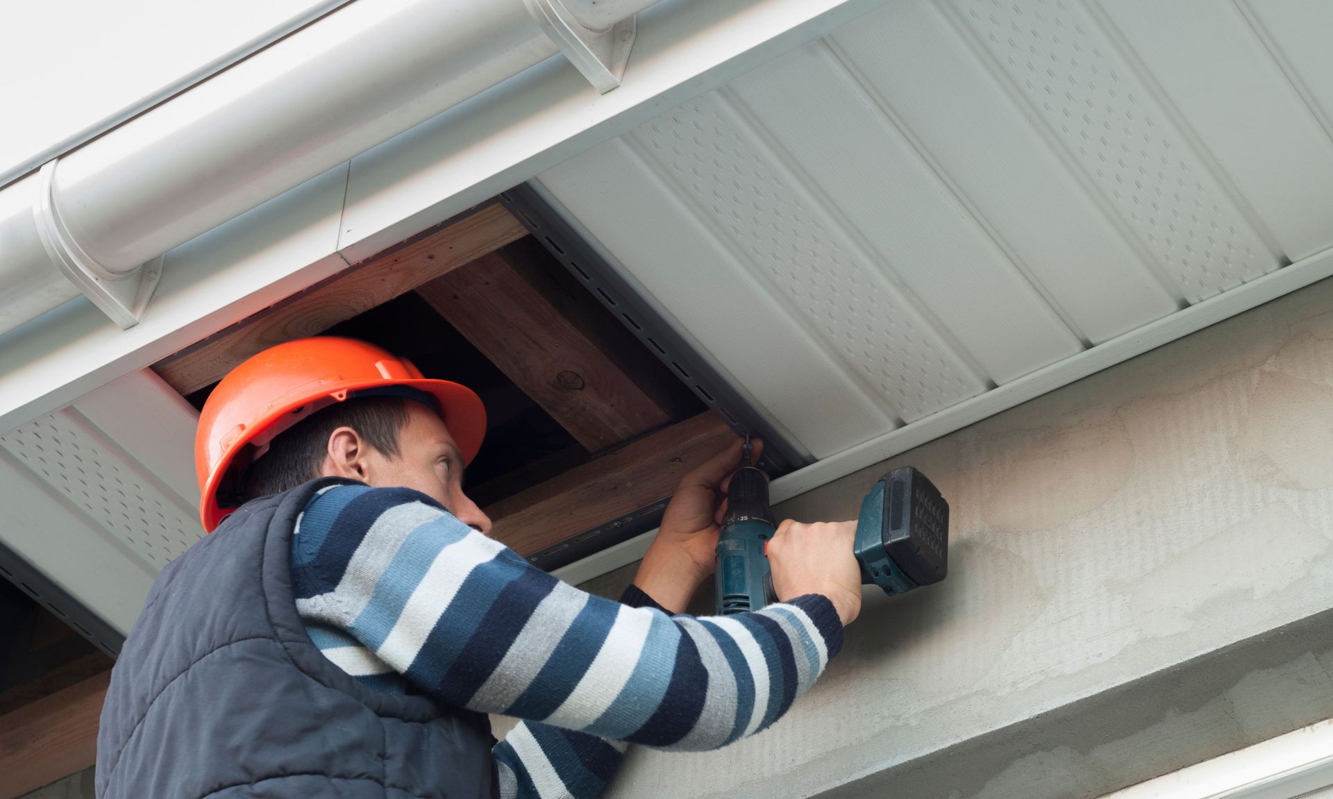 Man in hard hat uses a drill to work on a white soffit under a gutter.