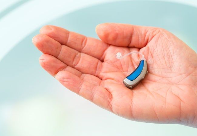 Open palm holding a small blue and silver hearing aid on a light background