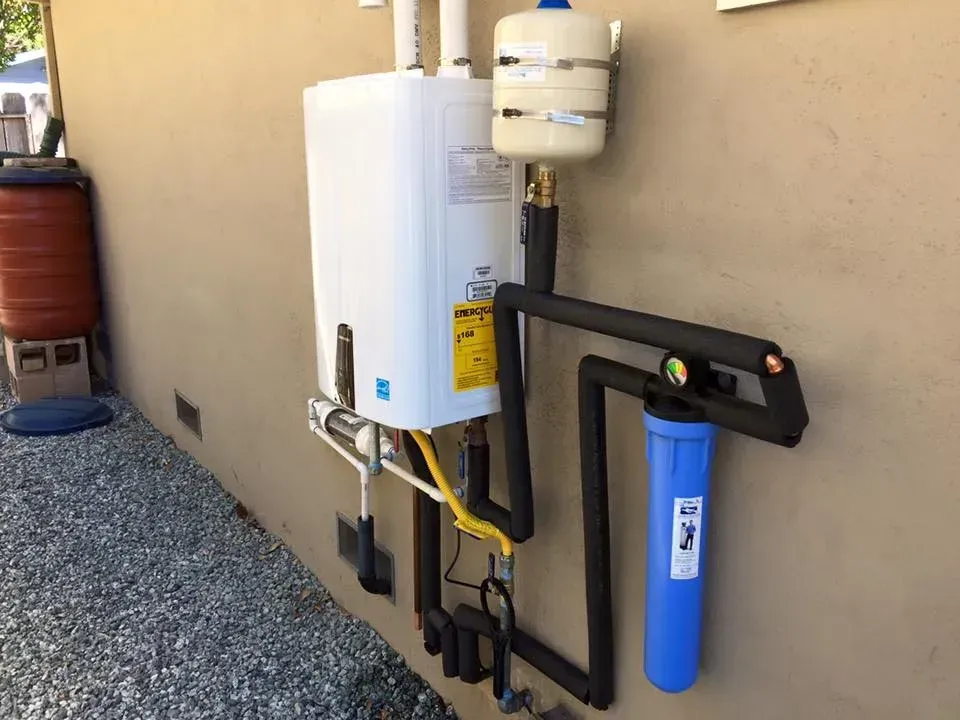 Wall-mounted water filtration system with white tank and blue filter beside a beige exterior wall.