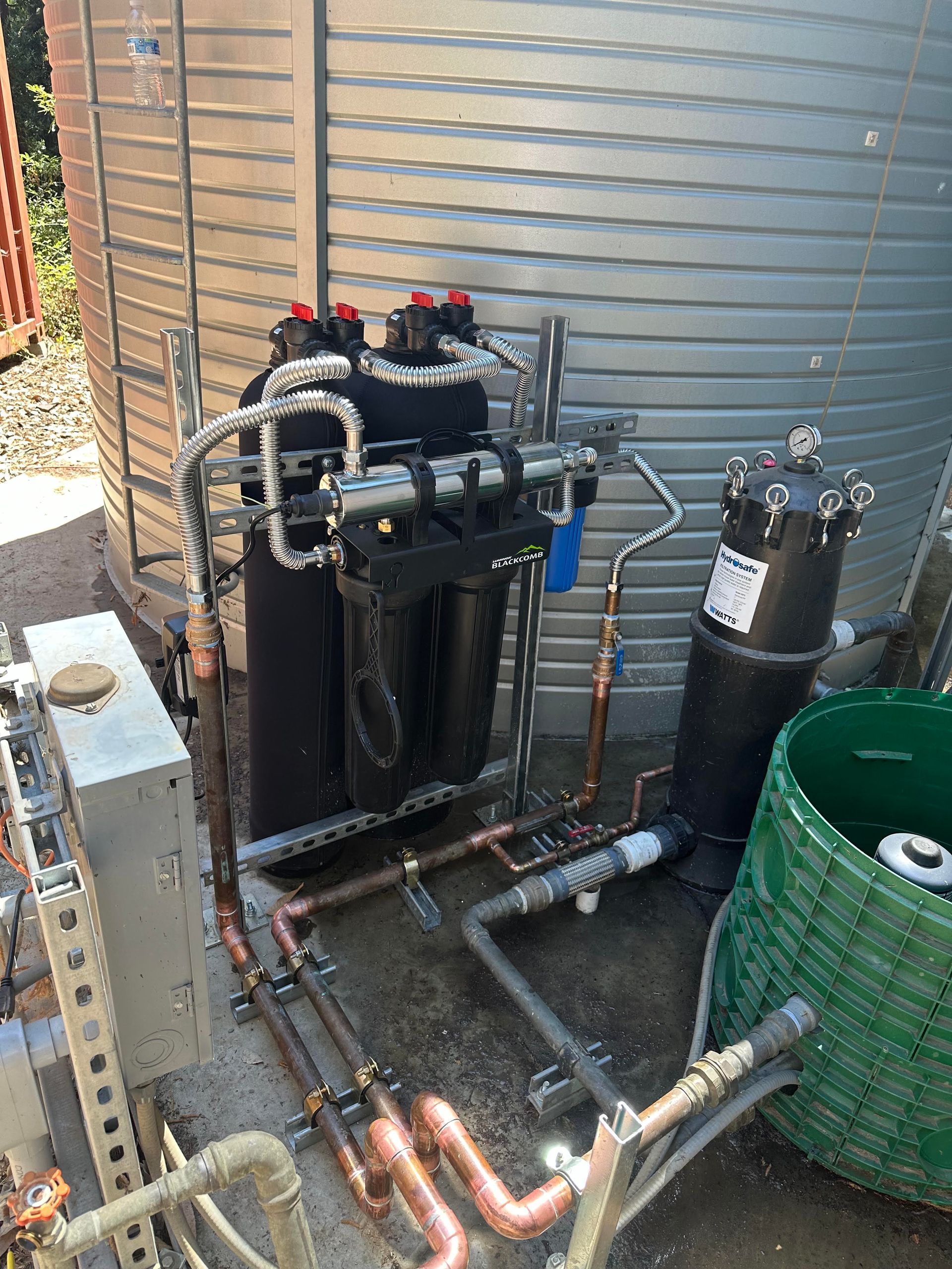 Outdoor mechanical equipment with black tanks, copper pipes, and hoses beside a corrugated metal wall