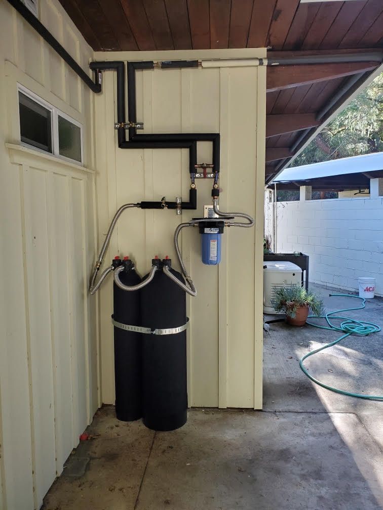 Two black water softener tanks beside a house wall with exposed plumbing and a blue filter system under a roof overhang