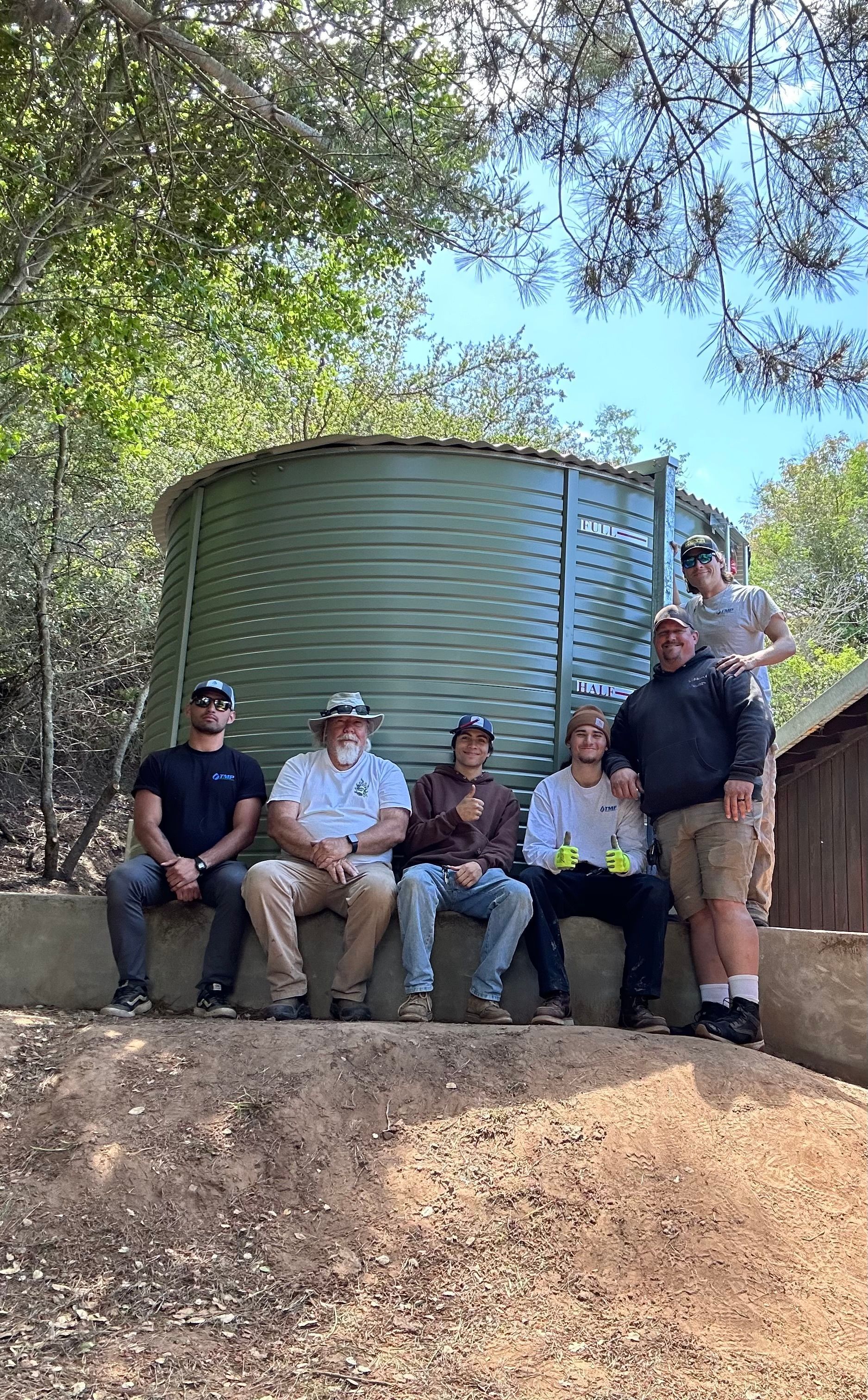 Five people posing in front of a large metal water tank under trees outdoors