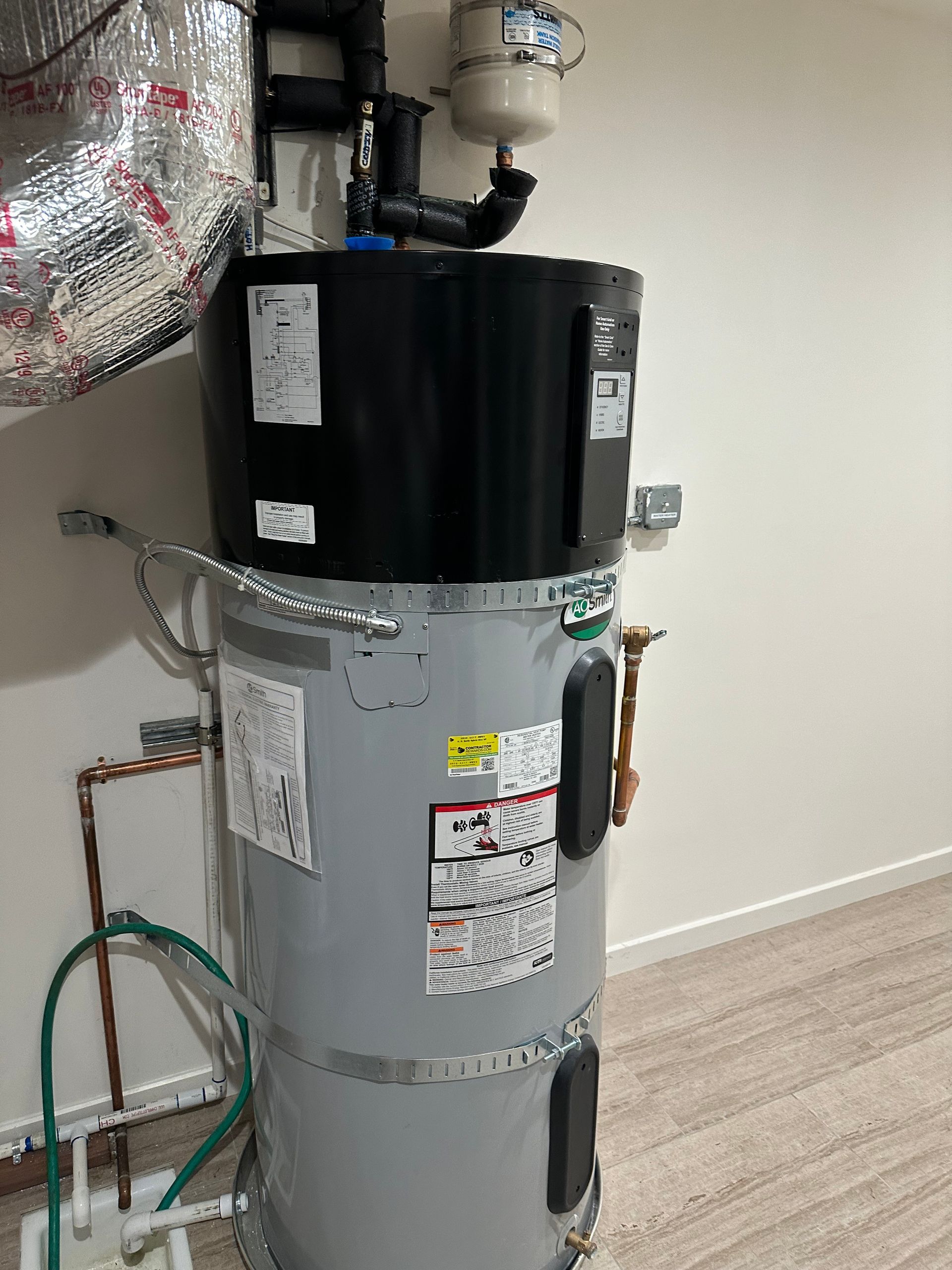 Water heater with black top and gray tank in a utility closet, with pipes and insulation above.