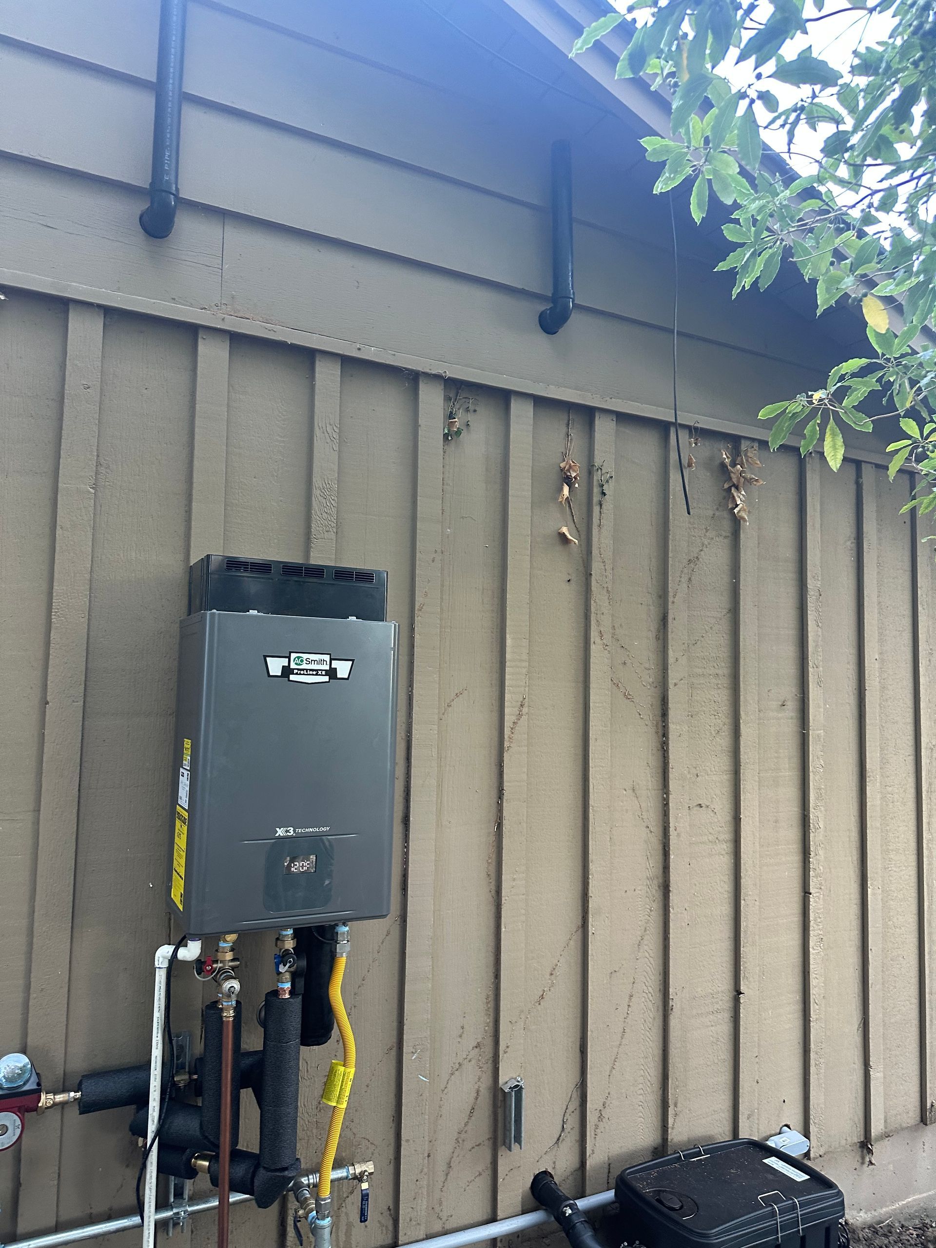 Wall-mounted gray utility box and pipes on a tan exterior wall under roof eaves, with tree leaves at right.