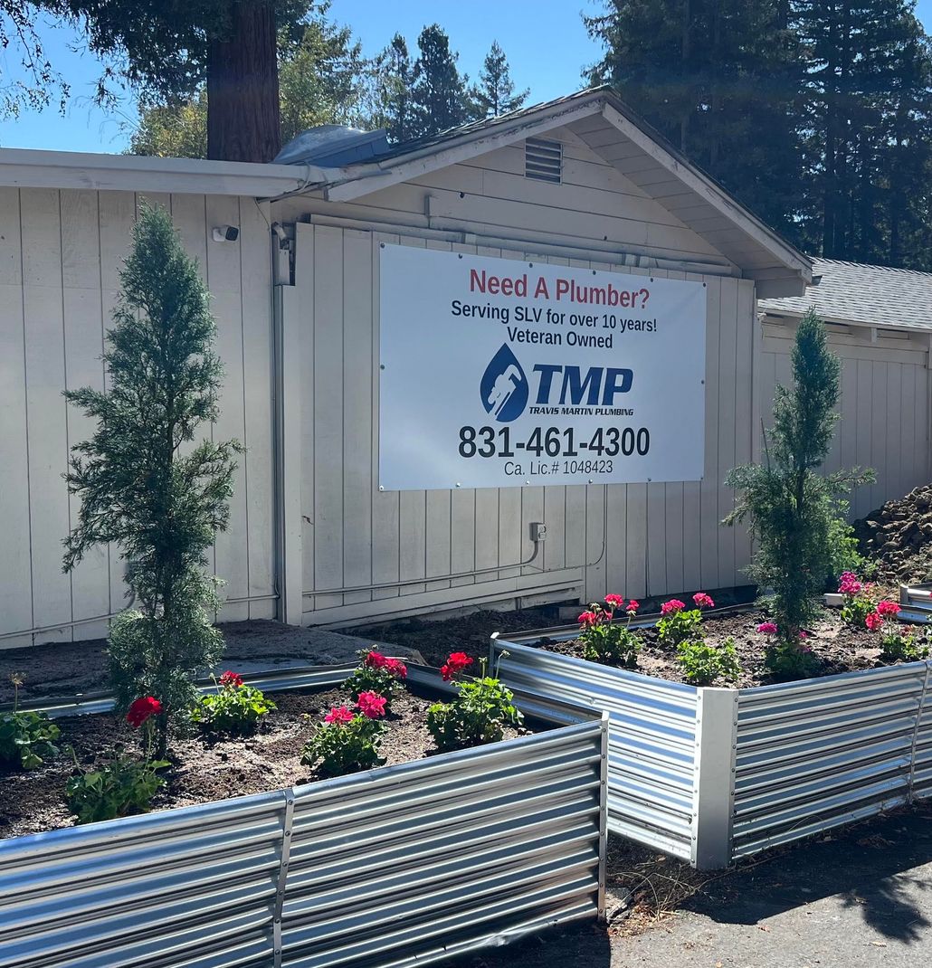 Plumber service banner on a beige shed, with flower beds and tall trees in the background.