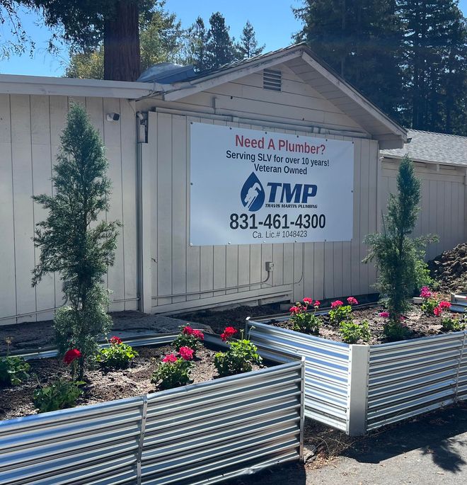 Plumbing service sign on a beige shed beside flower planters and trees in bright daylight