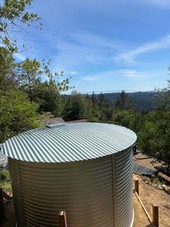 Large corrugated metal water tank beside trees, overlooking a distant hillside under a blue sky