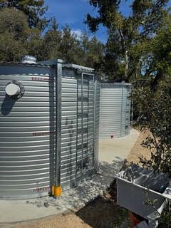 Large cylindrical metal water tanks beside a concrete pad and trees outdoors